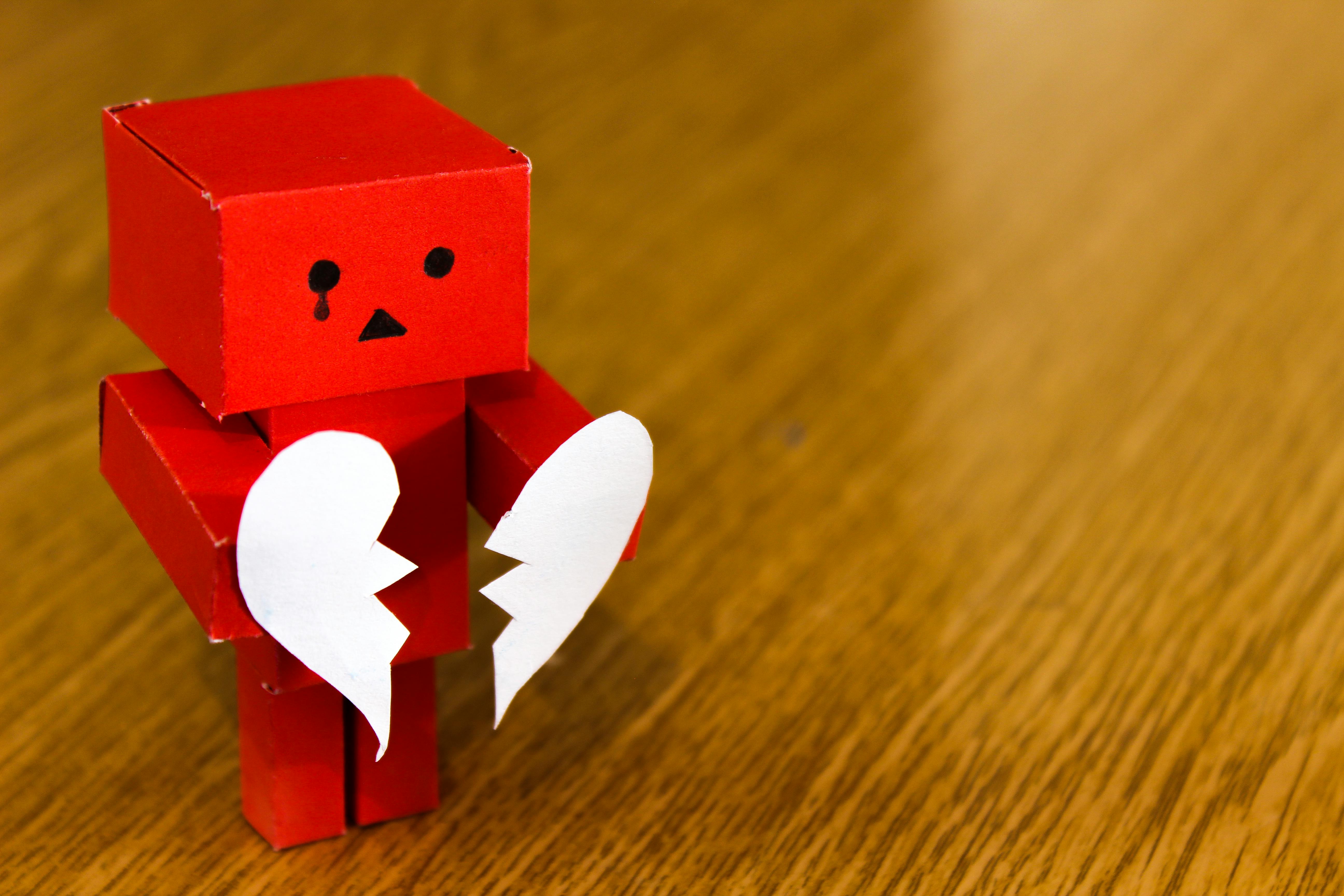A red, paper-made robot is standing on a table, holding a torn, white paper heart, one piece per hand.