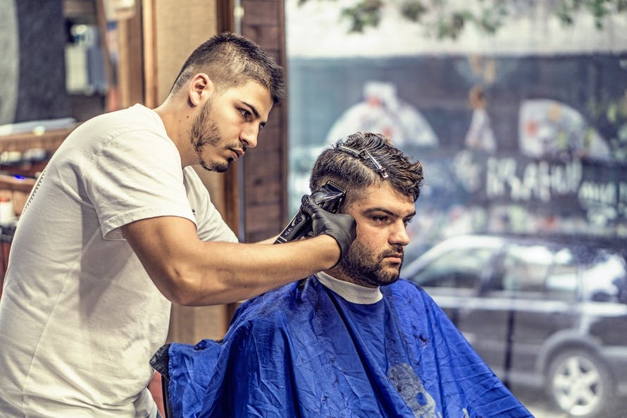 Barber in White Shirt Trimming Man\'s Hair in Blue Textile While Sitting Nearby Glass Window