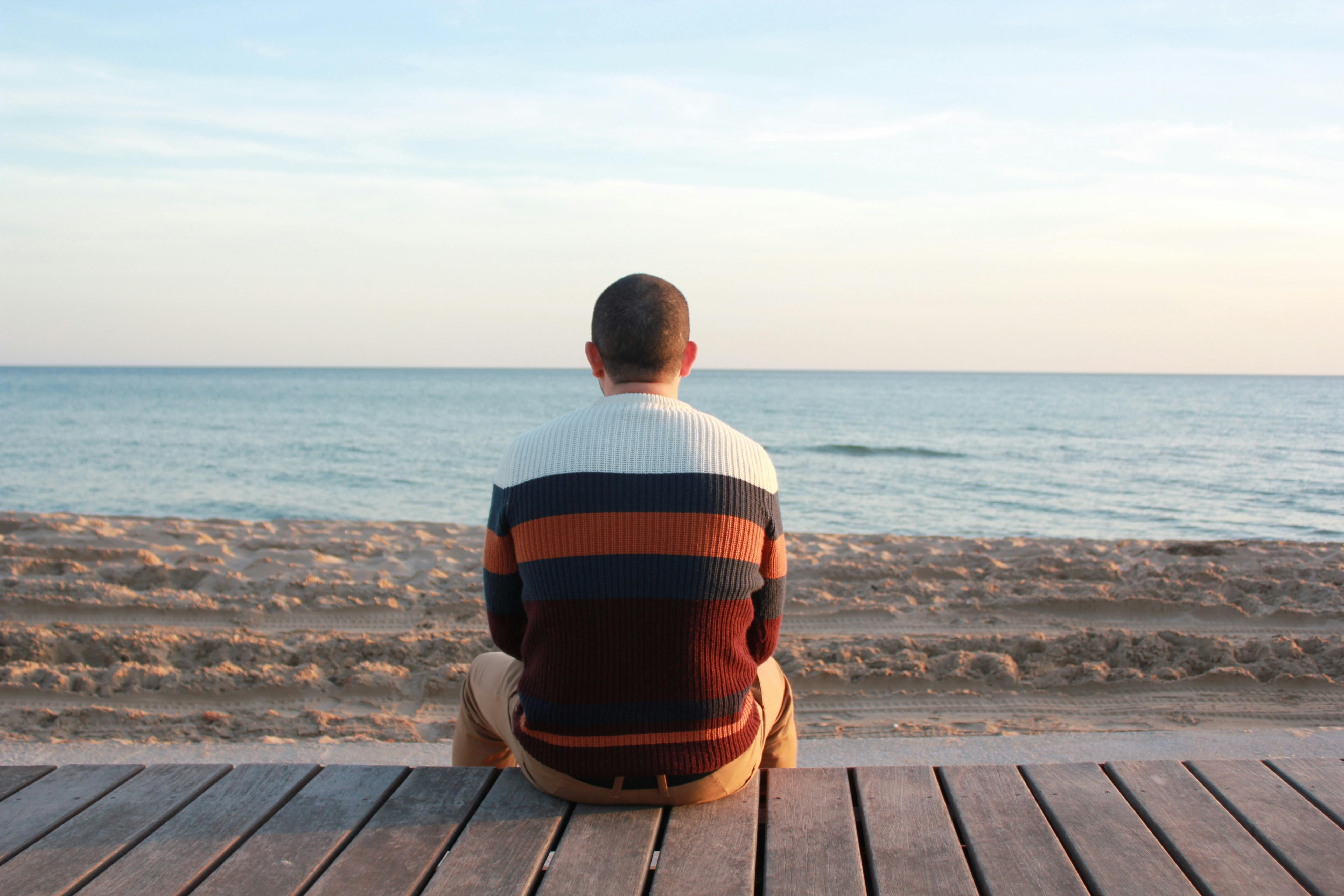 Man Sitting On Wooden Panel Facing In The Ocean · Free Stock Photo