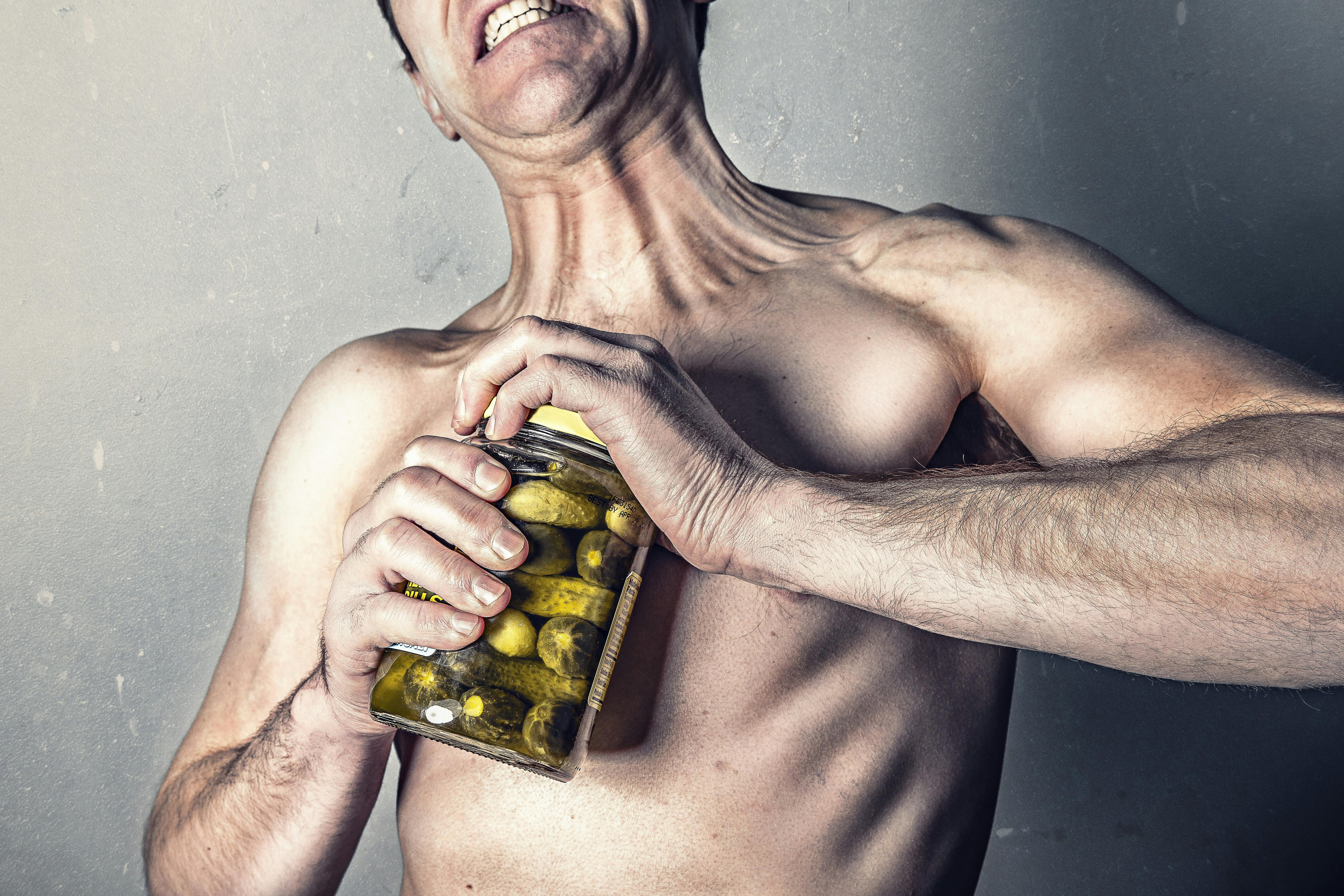 Shirtless man trying to open a jar of pickles.