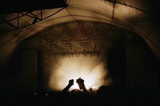 This photo shows a concert venue inside a huge tent. A crowd is gathering in front of the stage cheering. The stage itself is covered in fog and light. The photo has sepia colors.